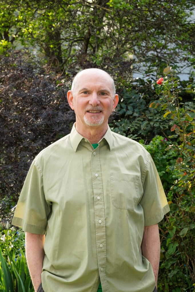 A photo of Mark Strong, wearing a sage green shirt, in front of foiliage.
